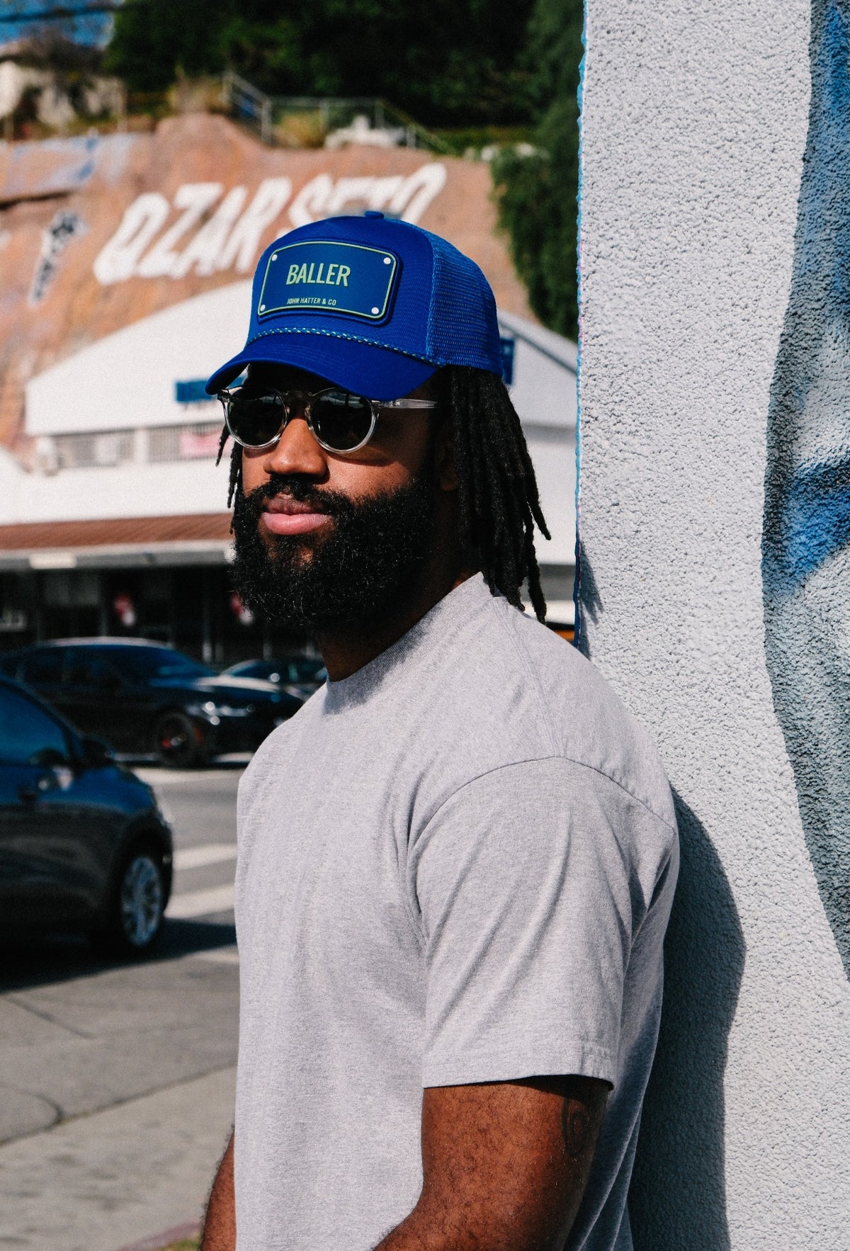 Man wearing a blue Baller John Hatter trucker cap, sunglasses and a grey T-shirt
